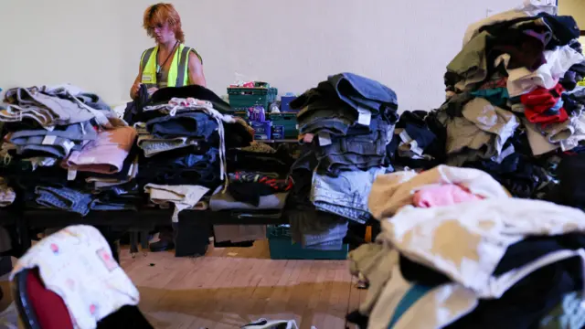 A person in a yellow and silver hi-vis jacket is sorting through piles and piles of clothes on a table
