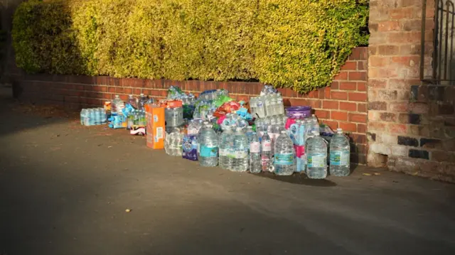 A pile of supplies, including large botles of water, next to a wall on a pavement.