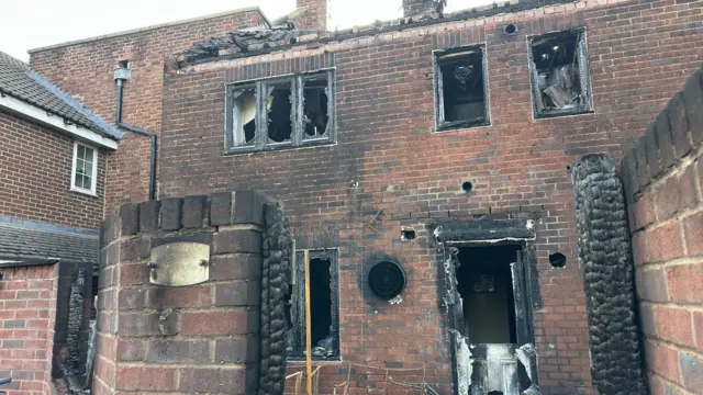 A house affected by fire. Most of the roof has gone and wooden beams can be seen. All of the windows are blackened and have exploded as well as the door
