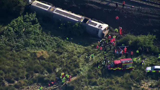 An aerial view of a train in a field on its side surrounded by emergency crews.