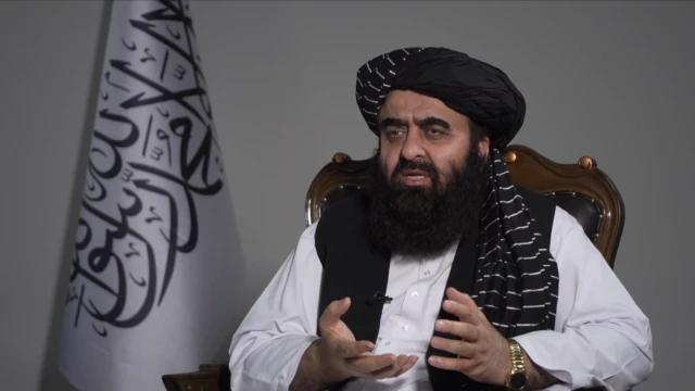 Amir Khan Muttaqi sits in a chair and speaks while articulating his hands. There is a white and black flag behind him.