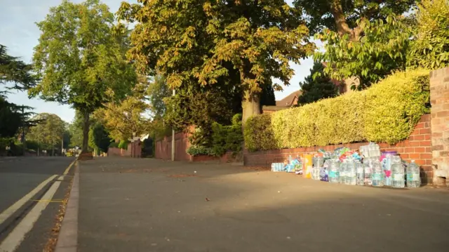 A pile of supplies, including large botles of water, next to a wall on a pavement.It is located on a quiet residential street with trees visible alongside.