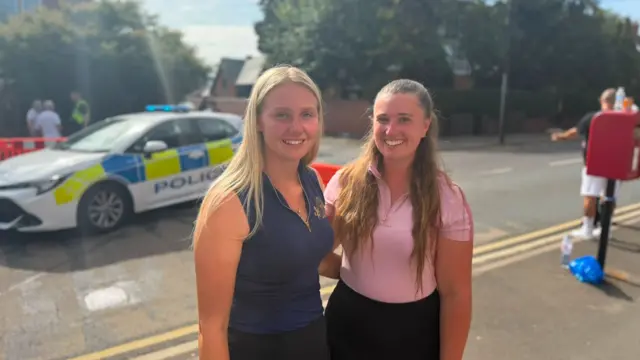 Izzy (left) and Abbie (right) standing together smiling for the cameras. Izzy has blonde hair and is wearing a navy blue top, Abby has a pink top on and dark trousers. Behind them is a police car and cordon on the road.