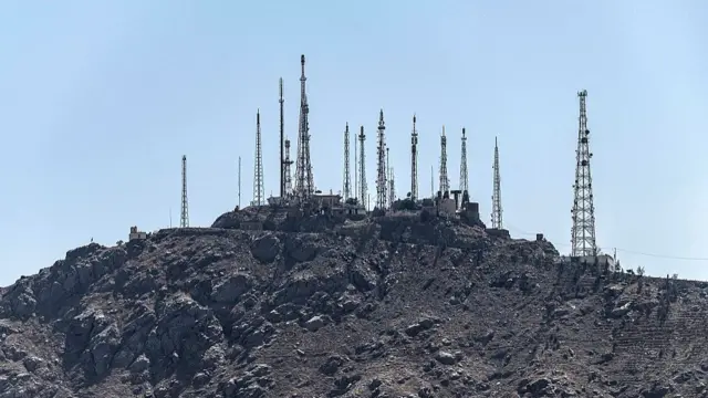 Several telecommunications towers seen on a hilltop against a light blue sky.