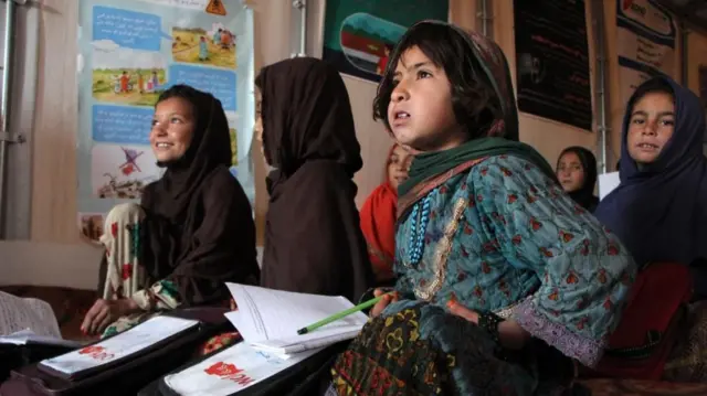 Half a dozen young girls wearing headscarves are shown sitting on the floor with lesson books and pencils.
