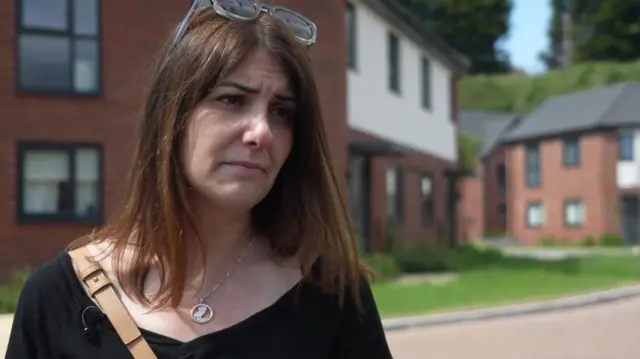 Cat Eccles stands in front of a row of houses on a sunny day. She has long brown hair and has her sunglasses on her head while she is wearing a black top with a light brown bag.