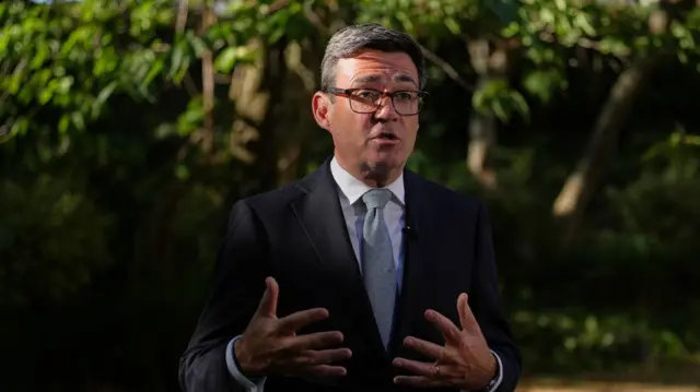 A man wearing glasses and a dark-colured suit is standing outdoors. He has a white shirt on and a light-coloured tie.