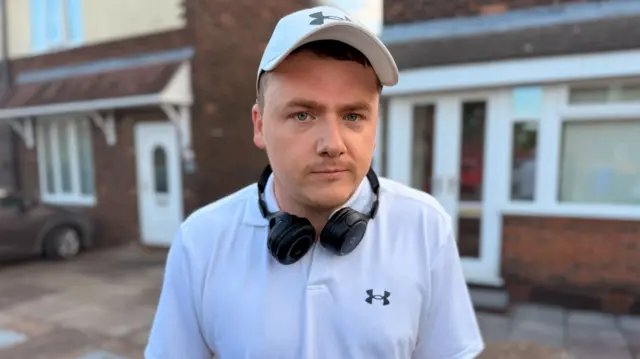 A man with a white hat, black headphones around his neck and a white t-shirt, stands outside the front of a house and looks at the camera.