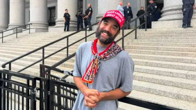 Kristopher Aadahl  stands outside court wearing a grey shirt with a neckerchief and a red hat saying Make America Great Again. He had a dark beard and is smiling at the camera.