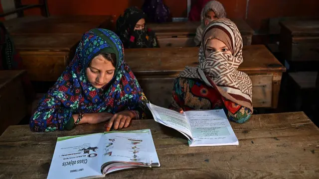 Two young girls sit at a school desk reading books. There is another pair of girls sitting behind them, with an empty desk in front of them.
