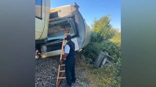 A man with a ladder helping people exit a train carriage on its side.