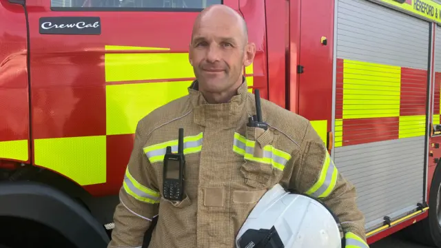 Alex Hustwayte - a bald man wearing a firefighter's outfit and holding a white helmet - smiles slightly as he stands next to a fire engine.