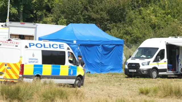 British Transport Police vehicles, investigation vans and a blue temporary tent stand in a field close to the railway near Lewes. Personnel are gathered around the temporary facilities being used during the investigation.