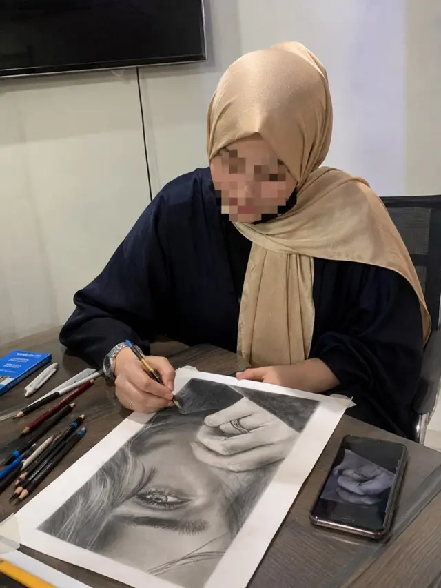Shakiba drawing a picture of a woman at a desk, with pencils on the table, and her face pixalated