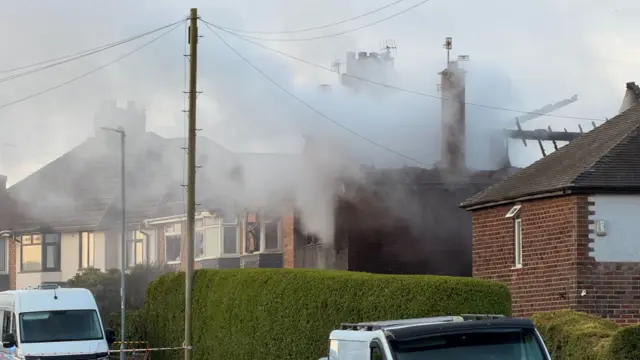 Smoke rises from a house that has been destroyed by a fire in a residential area. A couple of white vans can be seen parked on the road next to it.