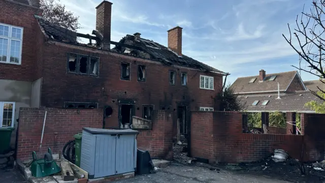A house charred and blackened by fire with windows blown out. Most of the roof is missing an there are beams exposed