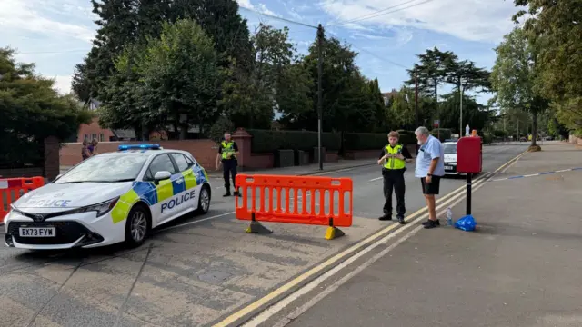 A general view of Norton Road in Stourbridge, a police car and big orange barriers are blocking the middle of the road. A police officer can be seen standing behind the car and a second officer is talking to a man in shorts and a blue shirt at the side of the road.