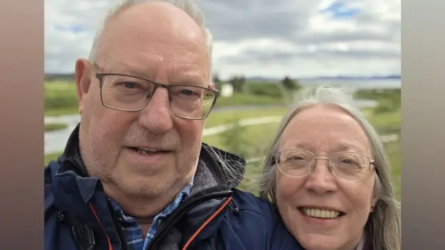 A woman with long grey hair and glasses smiles as she leans her head on the shoulder of a man with glasses and wearing a coat.