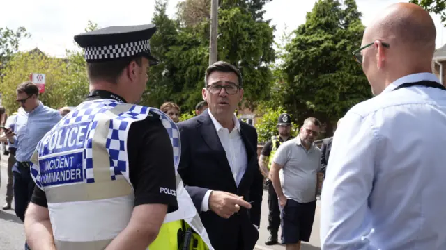 Andy Burnham in a black suit with a white shirt. He is talking to two police officers