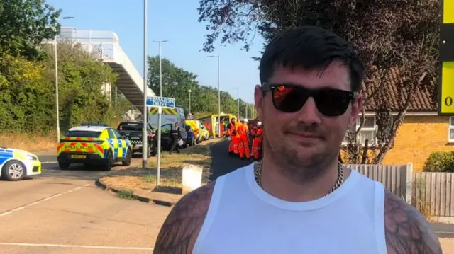 Aaron Poyser has dark hair and is wearing sunglasses and a white vest. He is standing in front of emergency crews at the scene.