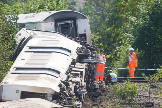 Two men in orange high vis and a police officer standing by train carriages on their side.