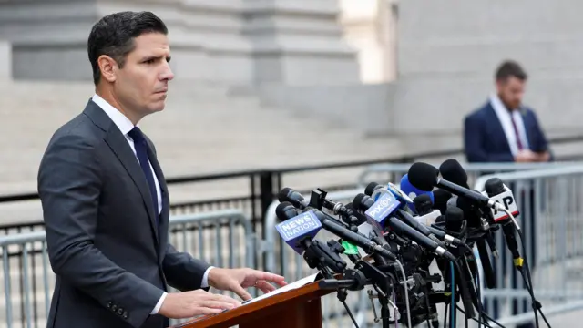 A man wearing a dark suit and a white shirt with a tie stands at a podium with microphones in front of him