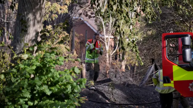 A firefighter stands on charred ground in a wooded area, with a building visible through the trees behind him. He is speaking on the phone as he walks towards a waiting colleague who is standing at the open door of a fire engine.