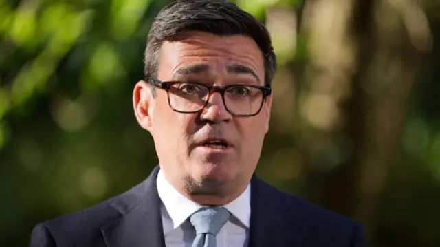 A man wearing glasses is standing outdoors wearing a white shirt with a light-coloured tie and a dark-coloured suit jacket. The image is a view of his head and shoulders.