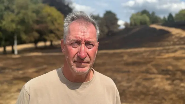Simon Marleym who has short grey hair and wears a beige T-shirt, stands in a field of brown grass