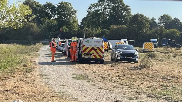 Vehicles and people in orange high-vis trousers and shirts in a field.