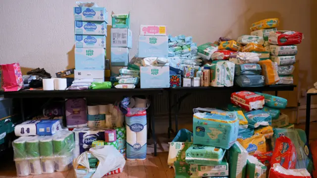 A huge pile of items for babies and young children on a table and on the floor around it. There are toilet rolls, nappies and baby wipes in colourful boxes