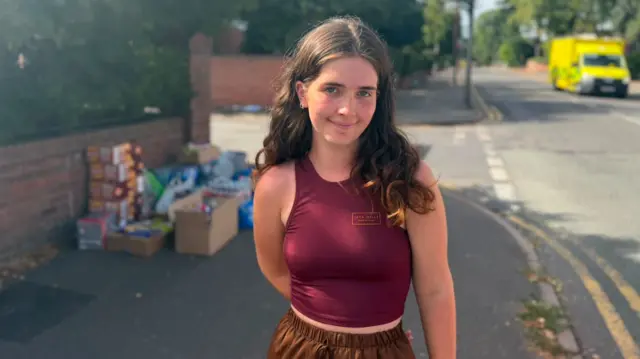 Olivia standing on the pavement smiling for the camera, she has brown trousers on and a purple vest top. She has dark hair and behind her you can see boxes of food and bottles of water that have been left for emergency services.