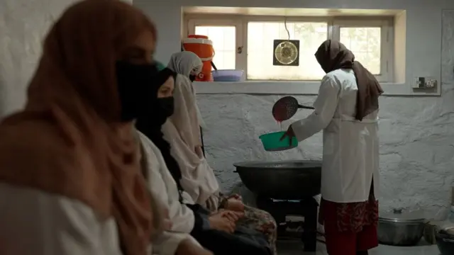 A woman using a large ladel to transfer jam from a large bowl into a smaller one, as other women sit in a line with their faces covered