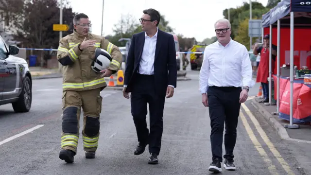 Three men are seen walking along a street. The man on the left of the image is wearing a firefighter's uniform, the man in the middle is wearing a dark-coloured suit with white shirt and open collar. The man on the right hand side is wearing dark trousers and a white shirt with open collar. All three are wearing glasses.