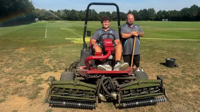 Thomas Belsey is sitting on a big motorised lawnmower with two giant blades at the front. Haydn Lawrance is stood to his right leaning on a big wooden stick. Behind them is a green lush football pitch with a hosepipe going across the pitch and water coming from it.