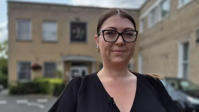 With brown hair tied back and large black glasses wearing a black v-neck top. She is standing outside a light bricked building in a car park that is out of focus.