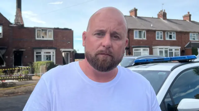 A bald man with a beard and wearing a white shirt, stands in the road looking sad. A house, just behind a tree behind him, has been damaged by fire and is missing most of its roof.