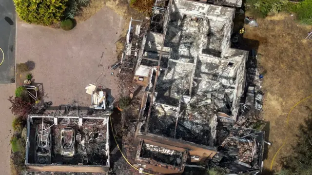 An aerial picture of a house completely destroyed by a fire. There is no roof and everything is either charred and blackened or covered in white debris. A small garage next to it is also destroyed, with two cars inside that are also in pieces