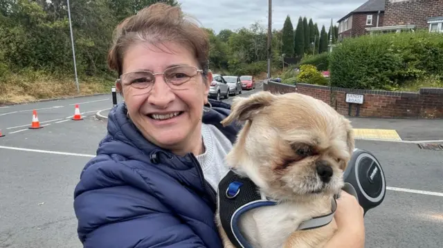 A woman in a navy puffer and glasses hugs a small beige dog in a harness and lead as she stands on a road with traffic cones behind her. She has short brown hair.