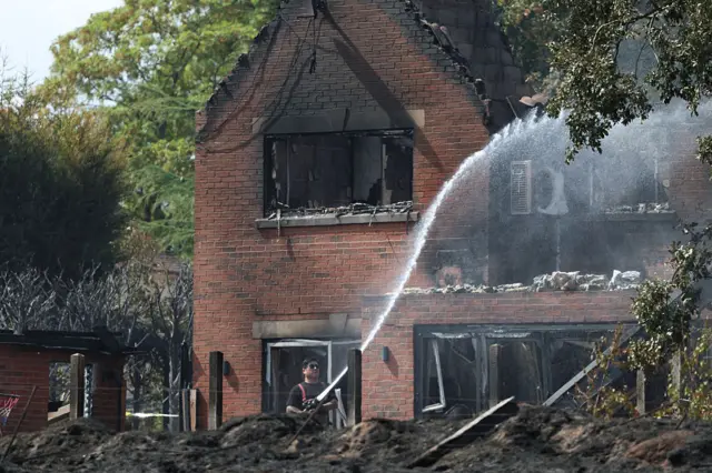 A lone firefighter in uniform (black t-shirt; red suspenders; and uniform trousers) douses a tree with water using a water pump. He's standing outside a red-bricked family home partially destroyed by fire, with charring marks around damaged windows