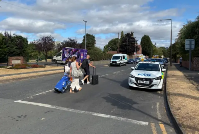 A man, a woman and a girl drag wheelie cases behind them as they cross a road with a police car parked at the end of it. he sky is blue with a few white clouds.