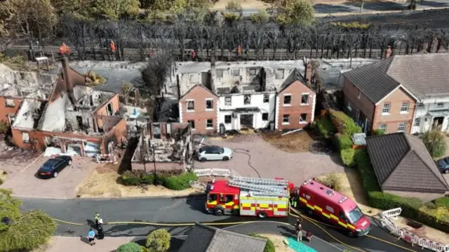 Drone footage shows an ariel shot of several homes which no longer have a roof or have fire damage. A fire engine and a red van can be seen at the front of the street and emergency workers can be seen walking through burnt trees behind the homes