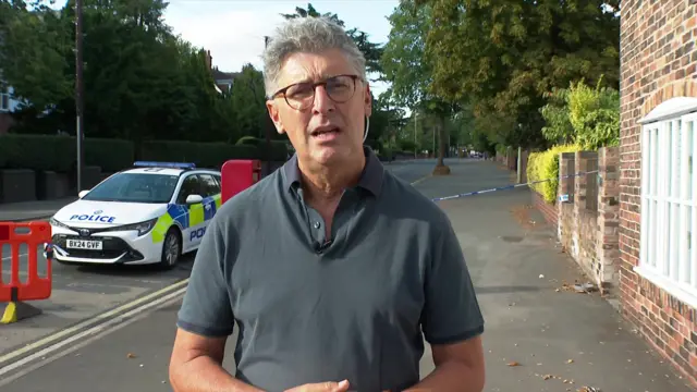 The Bishop of Dudles (middle-aged man with grey hair and black glasses) stands in the middle of a pavement looking into the camera. He's wearing a dark grey polo shirt, there's a parked police car behind him to his right