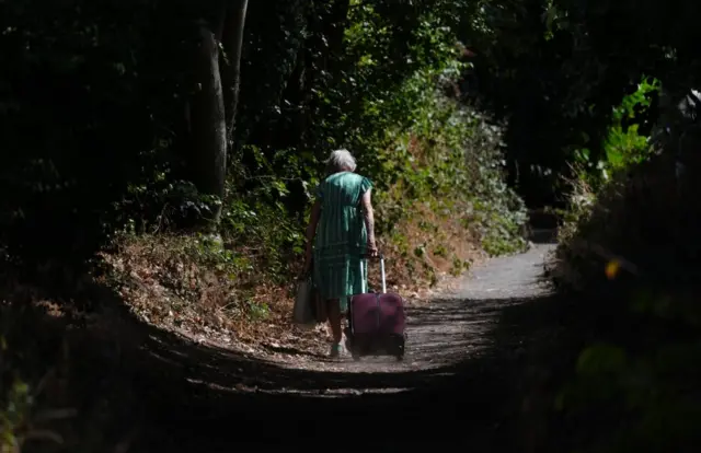 An elderly woman pulls a suitcase behind her as she walks down a country lane.