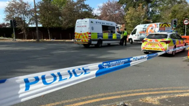 Police tapes stretches across a road, with police car and van seen beyond it. It is a sunny day. An orange cordon stands in front of the tape and a police officer in uniform and high-vis yellow vest stands by the van.