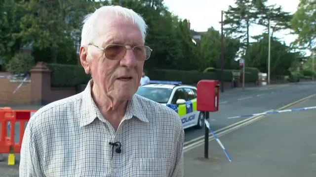 A man in a white shirt with blue and green checked pattern stands on a road looking at someone beyond the camera. He has white hair and glasses. Behind him, police tapis wrapped around a red post box and a police car is parked up. An orange cordon can just be seen.