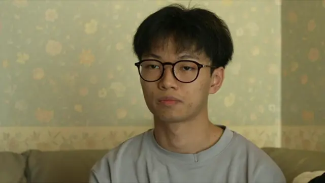 A teenage boy wearing a grey t shirt and black rimmed glasses sat on a sofa