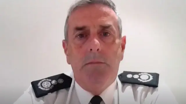 A male fire officer in white shirt and black tie with grey hair stares straight into the camera, with a serious expression.