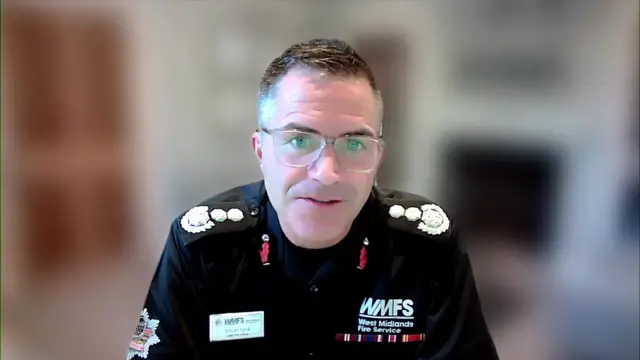 Chief Fire Officer Simon Tunhill in black uniform speaking to the screen during a remote interview. The background behind him is blurred