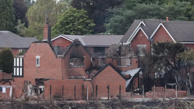 Wide shot of a residential area showing heavy fire damage to several red-bricked homes. One, at the centre of the shot, is missing the entire roof and has heavy scorch marks around windows